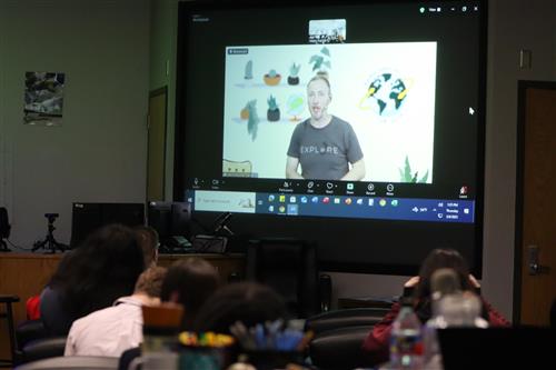  a class watching a projector in a classroom with a man speaking on it 